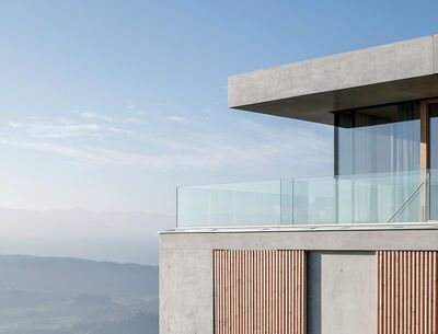 Modernes Einfamilienhaus mit Betonfassade, großen Fenstern und Glasgeländer auf einer Anhöhe mit weitem Blick über die Landschaft.