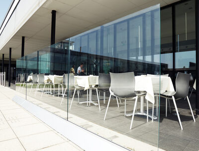 Glasabschirmung auf einer modernen Terrasse, die einen Außenbereich mit weißen Tischen und Stühlen abgrenzt.